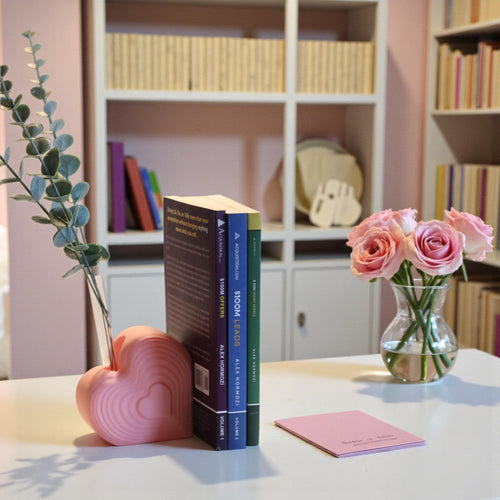 Pink heart bookend that doubles as a test tube vase, styled on a desk with books; romantic shelf decor and Valentine’s gift for book lovers.