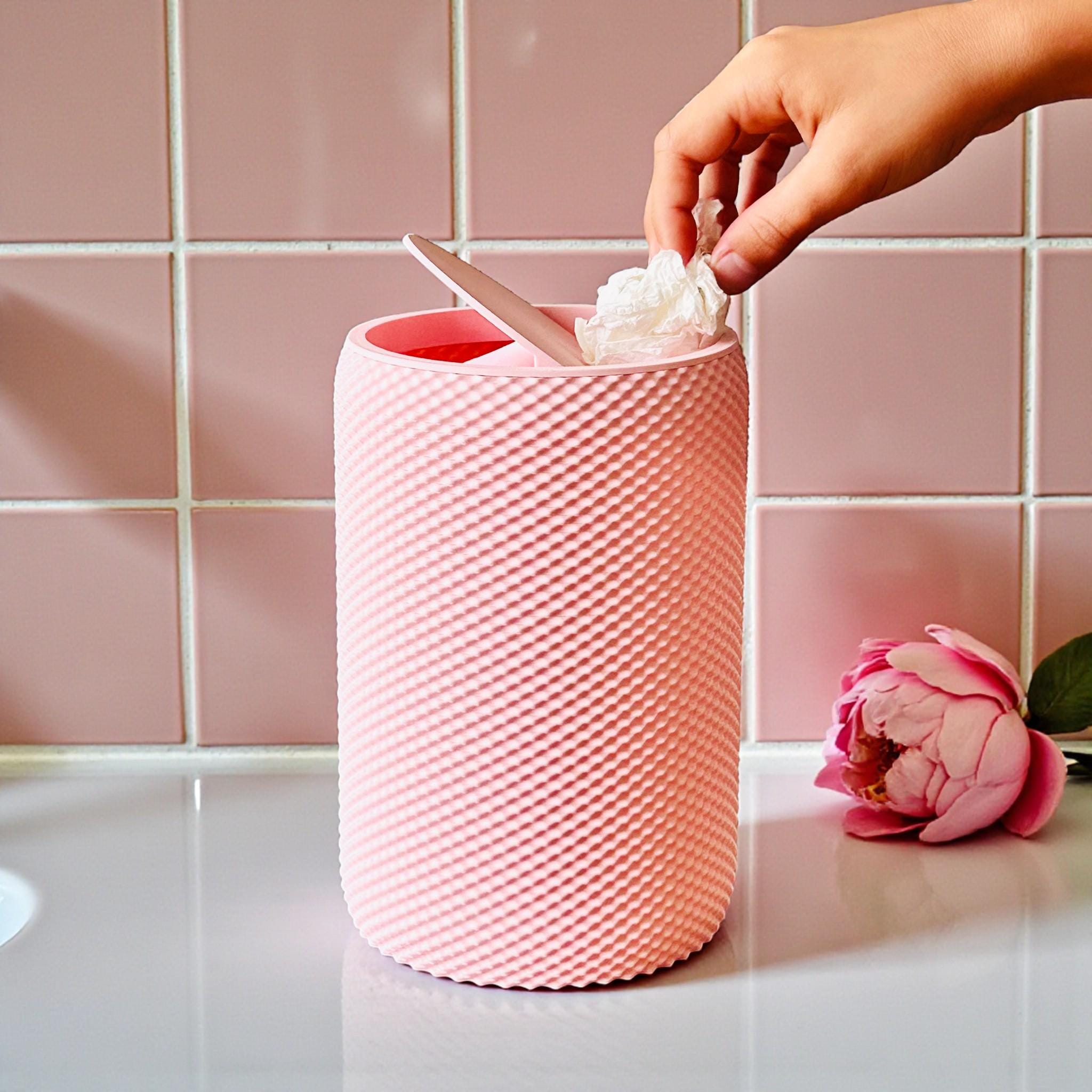 Pink textured wastebasket with a hand placing a tissue inside, against a pink tiled wall.
