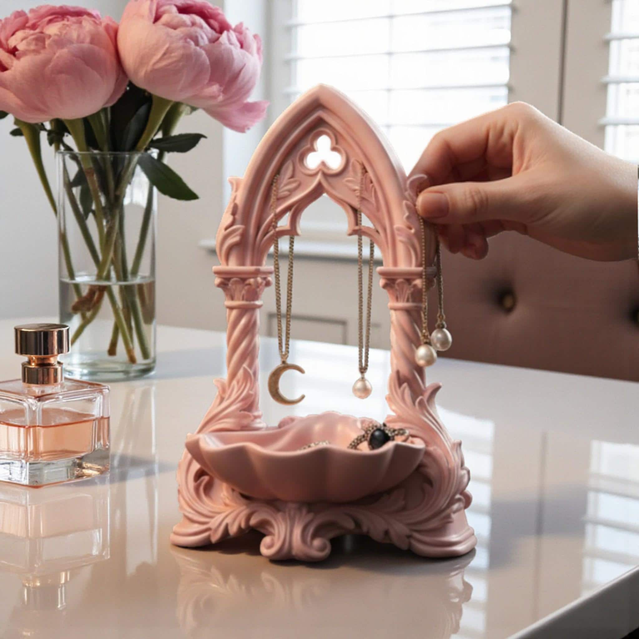 May include: A pink jewellery organiser with an arched design, displaying gold necklaces and earrings. A small dish at the base holds extra jewellery. The organiser sits on a dressing table with a mirror, perfume, and a vase of pink flowers.