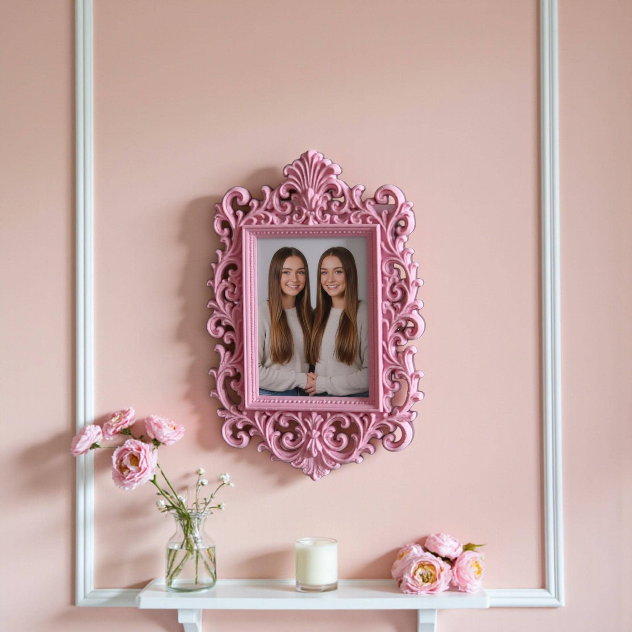 May include: A decorative pink picture frame with an elaborate, swirling design. The frame contains a photograph of a woman and child in a park. The frame is on a white shelf, with pink flowers and a candle.
