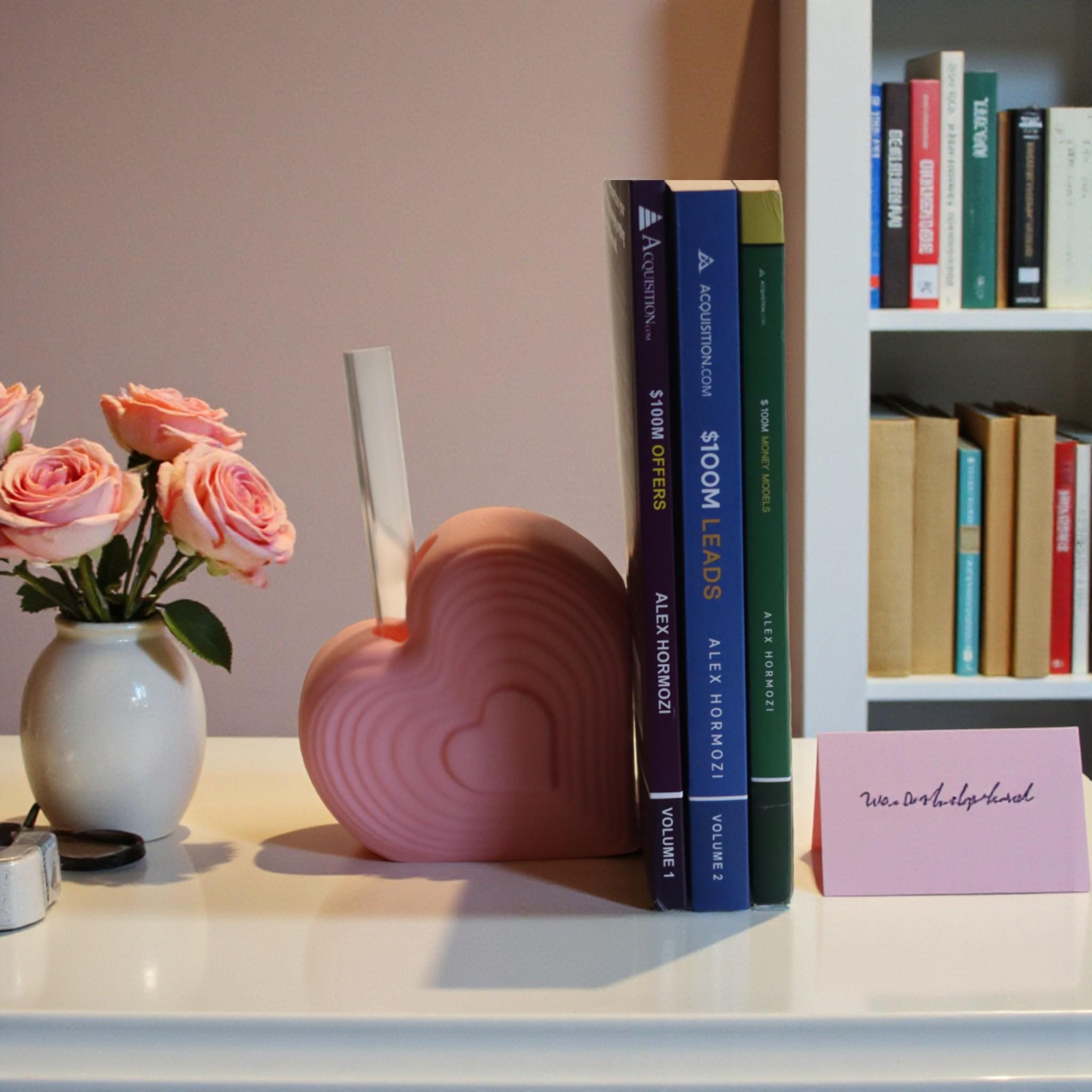 Pink heart bookend that doubles as a test tube vase, styled on a desk with books; romantic shelf decor and Valentine’s gift for book lovers.
