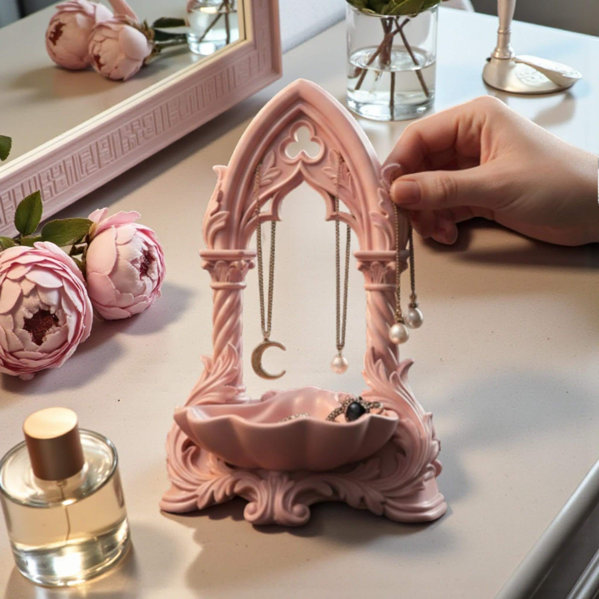May include: A pink jewellery organiser with an arched design, displaying gold necklaces and earrings. A small dish at the base holds extra jewellery. The organiser sits on a dressing table with a mirror, perfume, and a vase of pink flowers.