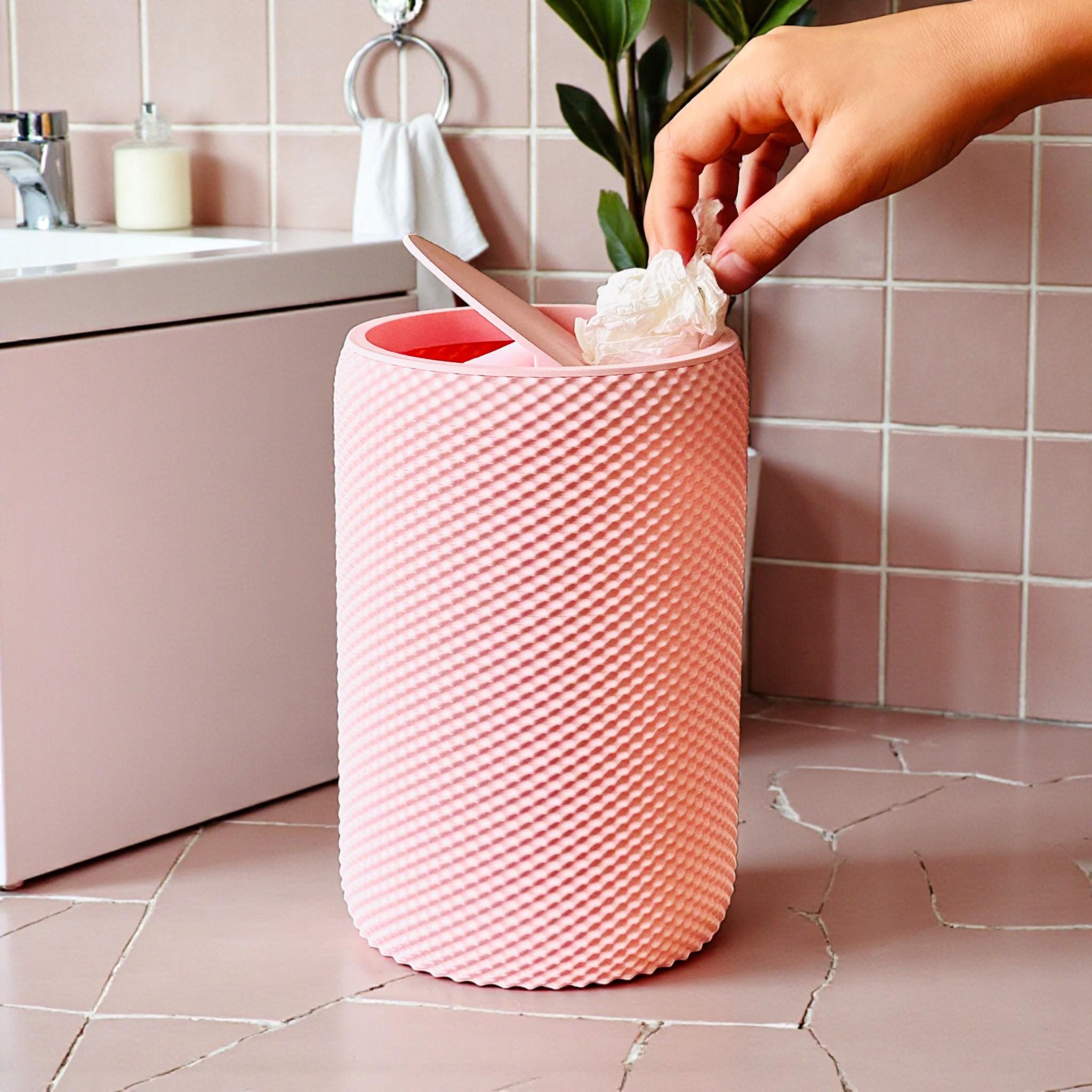 Pink textured wastebasket with a hand placing a white napkin inside in a bathroom setting.