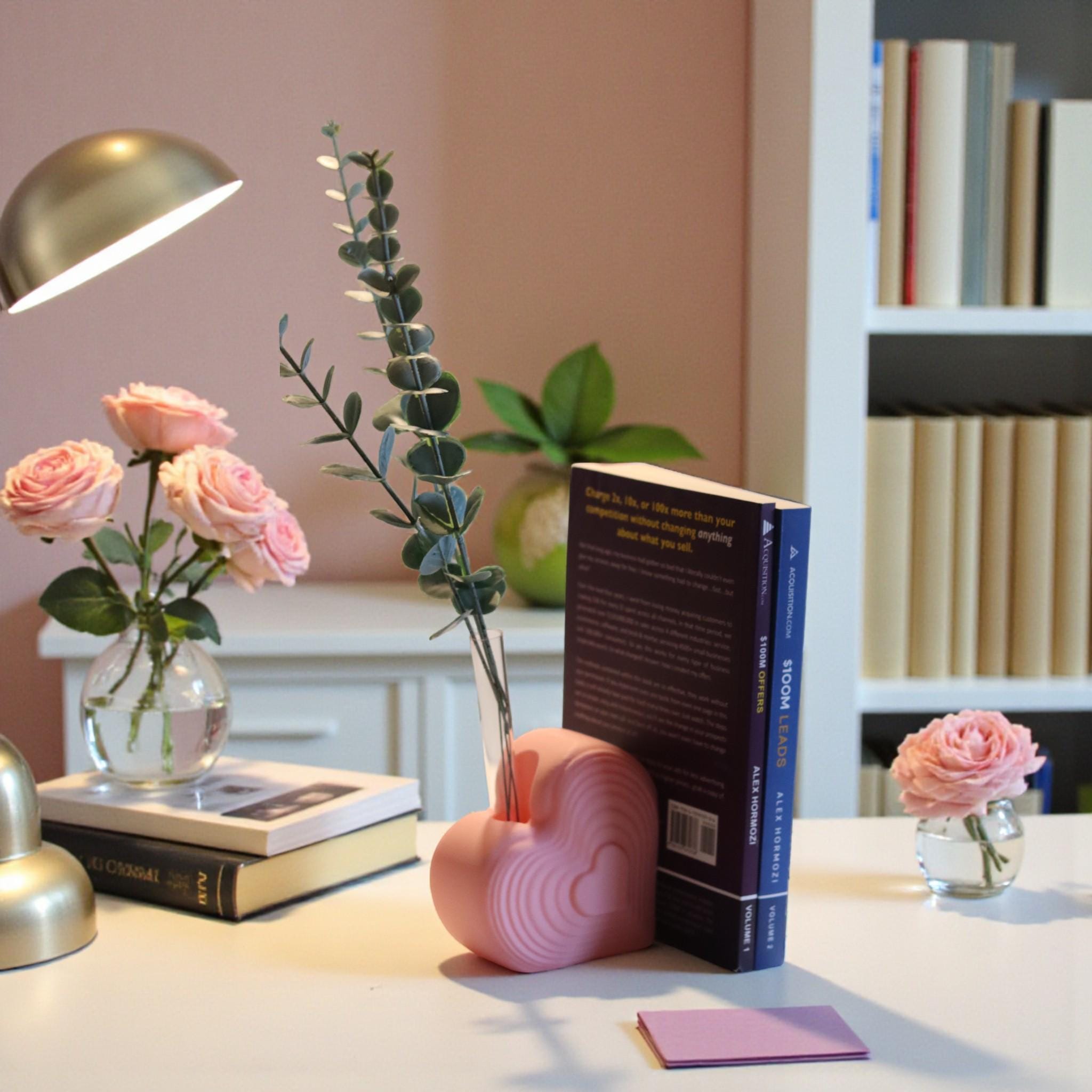 Pink heart bookend that doubles as a test tube vase, styled on a desk with books; romantic shelf decor and Valentine’s gift for book lovers.