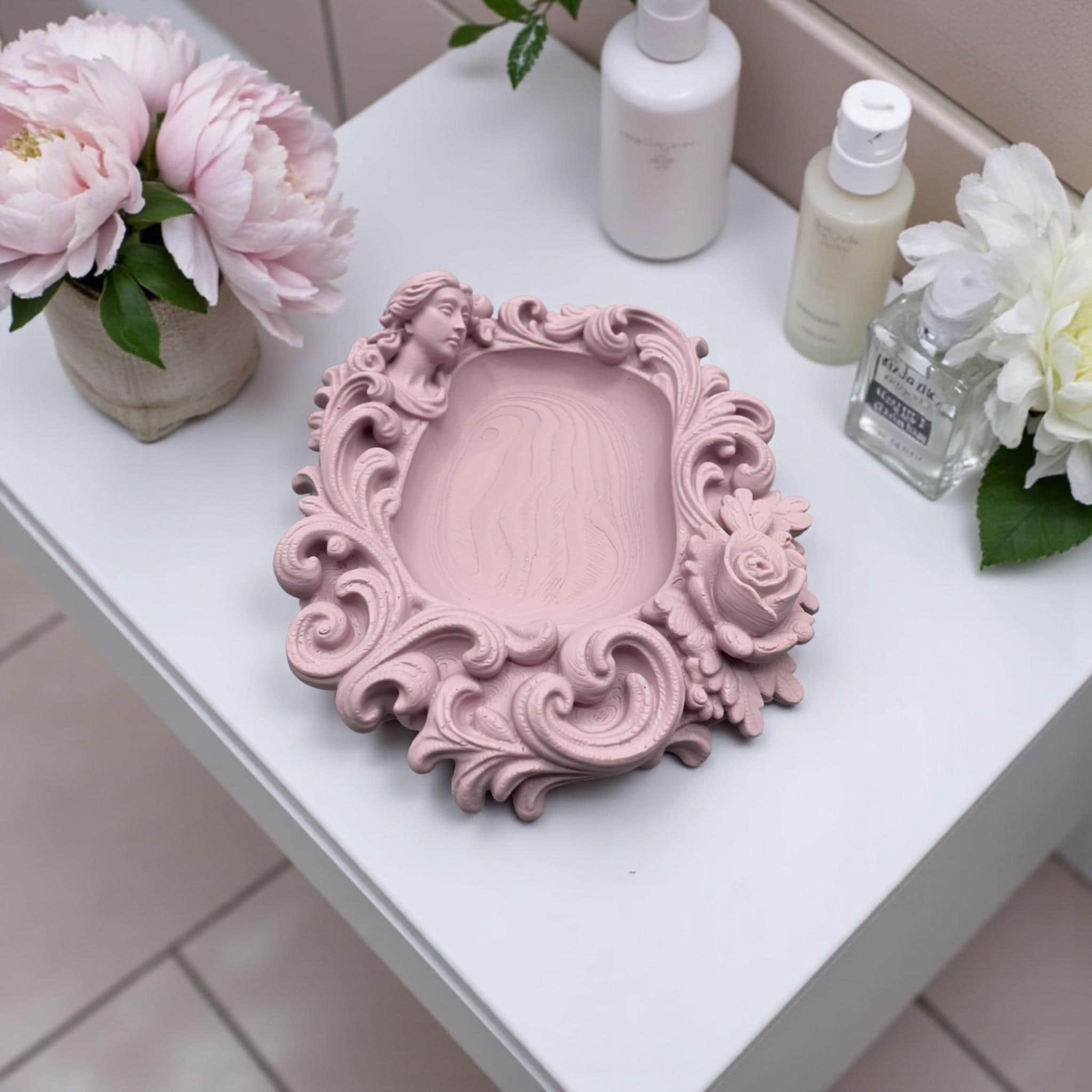 Pink rococo-style woman and rose catchall tray used as a jewelry dish, holding a gold ring, pearl earrings, and a necklace on a vanity surface.