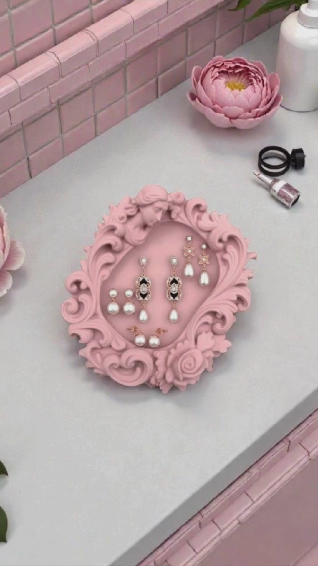 Pink rococo-style woman and rose catchall tray used as a jewelry dish, holding a gold ring, pearl earrings, and a necklace on a vanity surface.